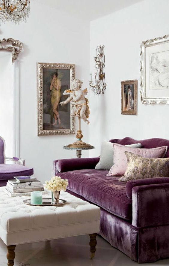 a refined living room with a mauve velvet sofa and pillows, a mauve chair, a creamy ottoman, some vintage art and a crystal chandelier