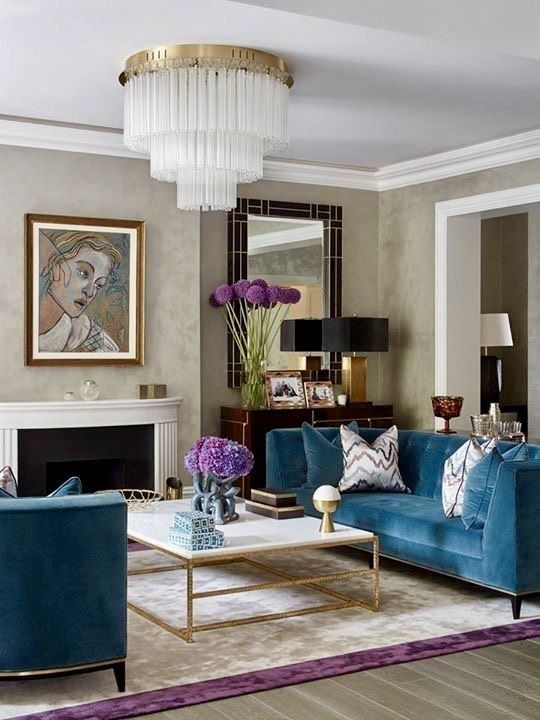 a sophisticated living room with a fireplace, blue velvet sofas, a coffee table, a statement chandelier, a console table and some purple blooms