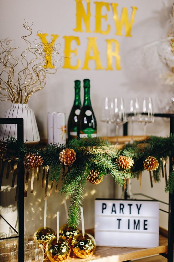 a NYE bar cart decorated with evergreens, pinecones, light branches, gold disco balls as glasses