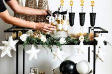 a NYE bar cart decorated with white stars, gold glitter cone trees, black glasses and black and white balloons