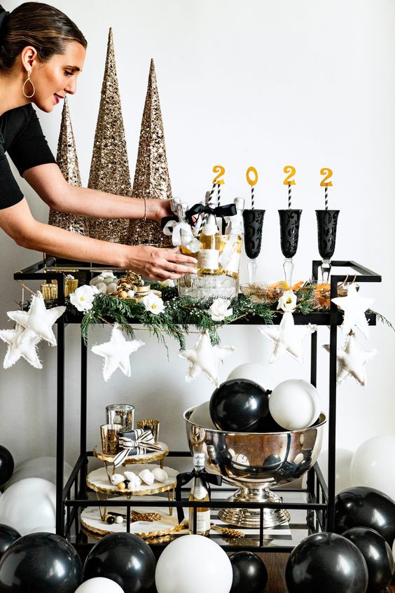 a NYE bar cart decorated with white stars, gold glitter cone trees, black glasses and black and white balloons