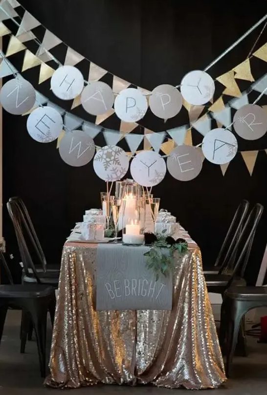 a NYE party tablescape with a gold sequin tablecloth, a neutral table runner and greenery, candles and sparkling garlands is a gorgeous space to celebrate NYE