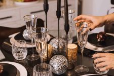 a NYE tablescape with black as a basic color, black candles and plates, disco balls, gold cutlery and gold-rimmed glasses