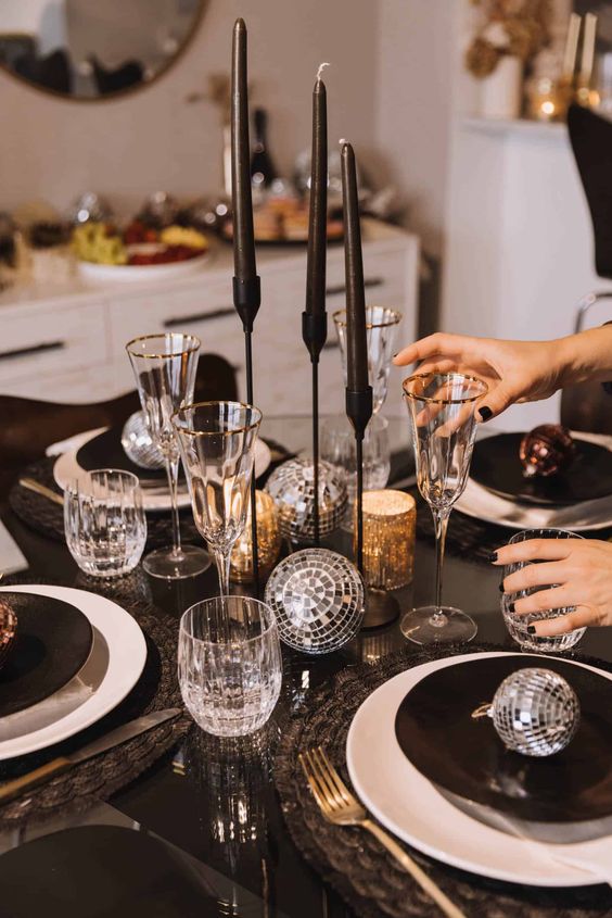 a NYE tablescape with black as a basic color, black candles and plates, disco balls, gold cutlery and gold-rimmed glasses