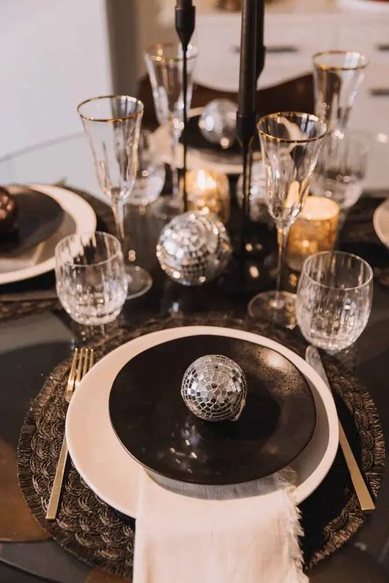 a black and silver NYE tablescape with black printed placemats, black and white plates, black candles and silver disco balls