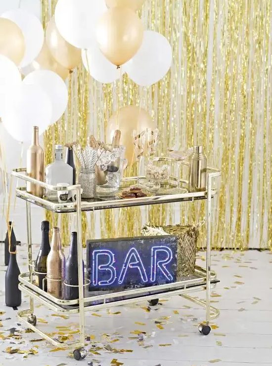 a brass bar cart with white and gold balloons, black and gold bottles and a neon sign