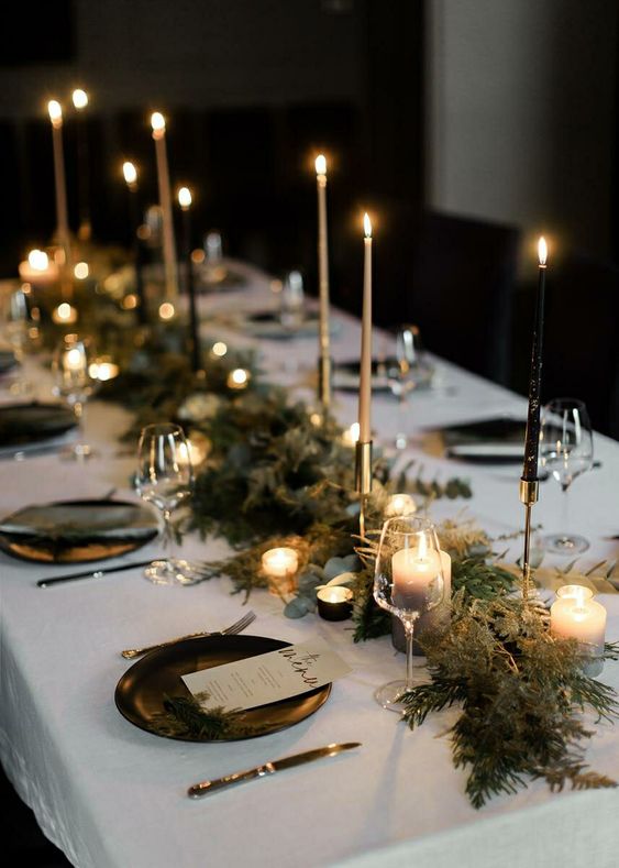 a chic NYE table with a green runner dotted with tall and thin and smaller candles, black plates and evergreens