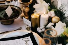 a chic NYE tablescape with a greenery runner, white blooms, cotton and gold ornaments, pillar candles, gold chargers and black napkins