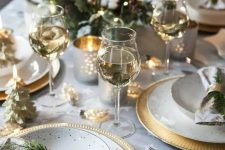 a glam NYE party table in neutrals, with golc chargers and gold candleholders, greenery, gilded Christmas trees and napkin rings