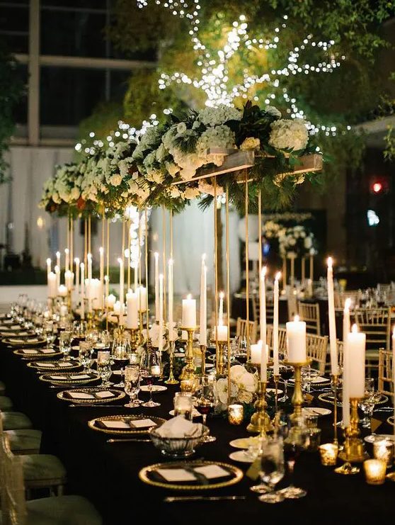 a gorgeous glam NYE tablescape with pillar and thin and tall candles, a tall centerpiece with white hydrangeas and greenery