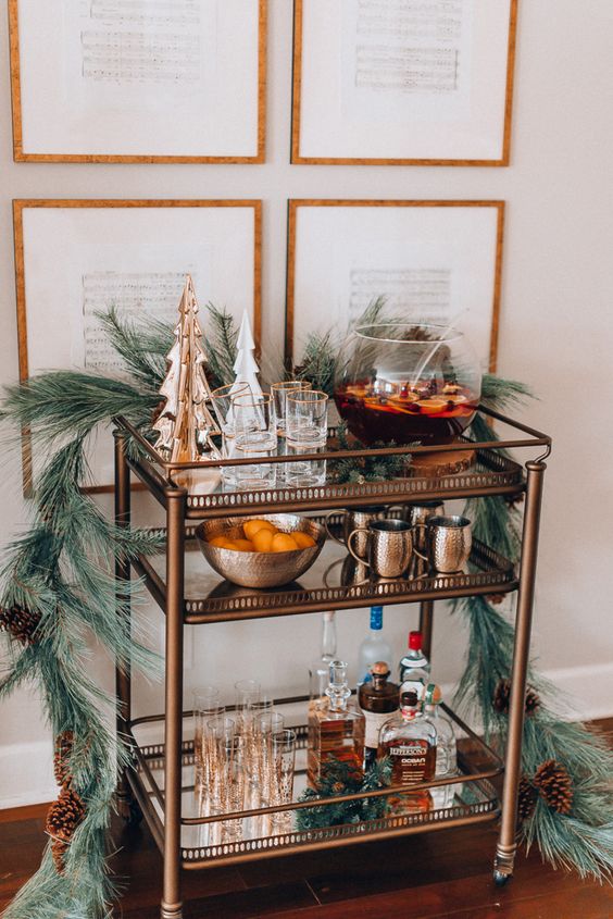 a holiday bar cart styled with an evergreen garland, tree figurines, pinecones and a tank with punch plus some barware