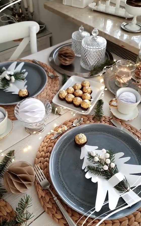 a modern Christmas tea party table with woven placemats, grey plates, sweets and candies, lights and evergreens