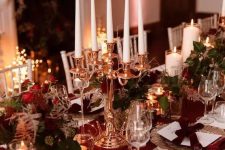 a refined NYE tablescape with a burgundy table runner and napkins, ornaments, greenery, red roses and a candelabra with tall and thin candles