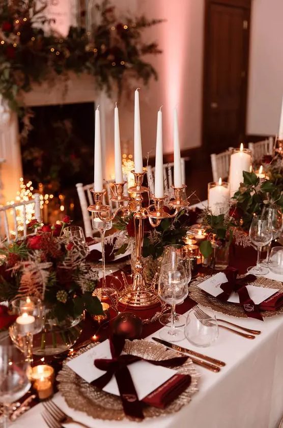 a refined NYE tablescape with a burgundy table runner and napkins, ornaments, greenery, red roses and a candelabra with tall and thin candles
