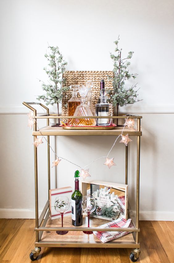 a simple holiday bar cart decorated with potted trees, a star light garland, a snowflake and some alcohol and glasses