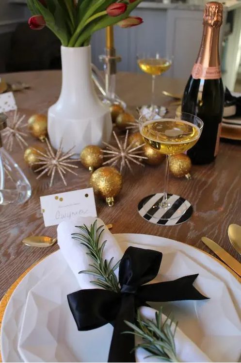a stylish NYE party tablescape with gold and copper ornaments, blooms, gold chargers and white porcelain, white napkins with a black bow