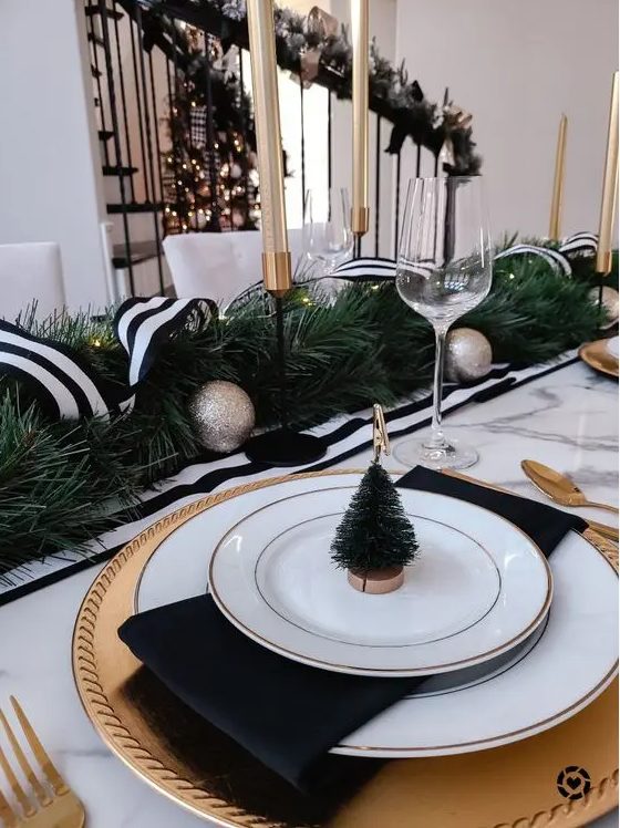 a stylish black and white NYE party table with gold chargers and white plates, black napkins, an evergreen runner with a ribbon and tall and thin candles