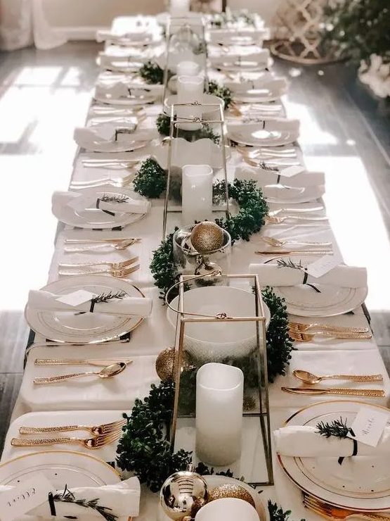 an elegant neutral New Year tablescape with white linens, gold cutlery, gold and gold glitter ornaments, a greenery garland and pillar candles