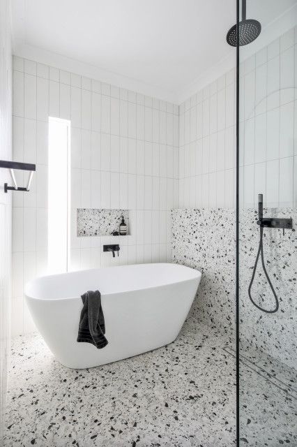 a black and white bathroom with white skinny tiles, white terrazzo tiles, a tub, a narrow window and black fixtures