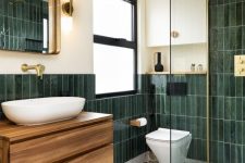 a bold modern bathroom with dark green skinny tiles and white terrazzo floor, a stained vanity, a mirror and gold fixtures