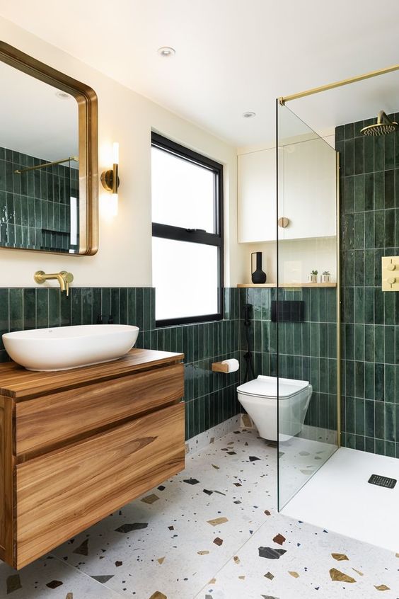a bold modern bathroom with dark green skinny tiles and white terrazzo floor, a stained vanity, a mirror and gold fixtures