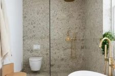 a cool modern bathroom clad with grey terrazzo, with a shower space, an oval tub, a wooden stool and some gold and brass fixtures