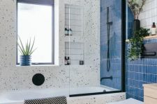 a cool modern bathroom with white terrazzo, a bold blue skinny tile wall, a tub with a window over it, a stool and some greenery