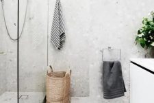 a grey bathroom completely done with terrazzo, with a white vanity and a basket and greenery to soften the look