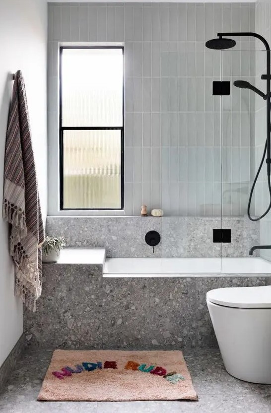 a modern bathroom done with grey terrazzo and grey skinny tile, black fixtures, a colorful rug and towel