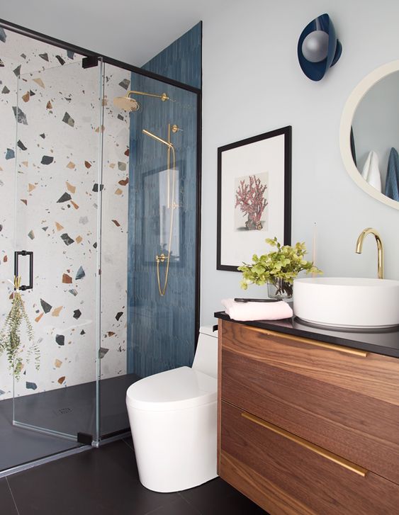 a modern bathroom with a terrazzo and navy tile wall, a shower space, a stained vanity, a round mirror and sink, a navy sconce