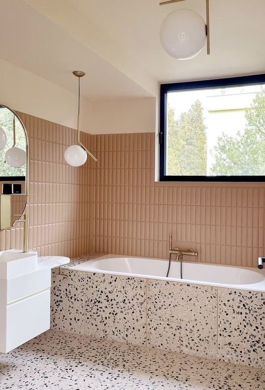 a modern bathroom with dusty pink skinny tiles, blush terrazzo, a white vanity, pendant lamps and a window