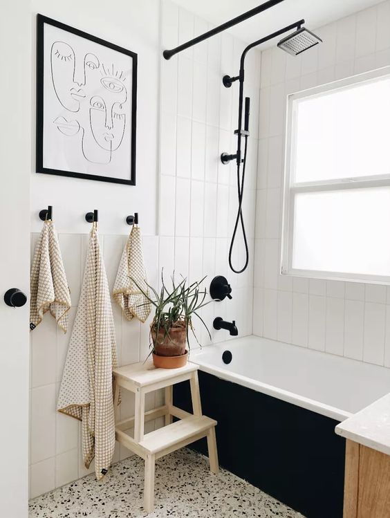 a modern black and white bathroom with white skinny tiles and a white terrazzo floor, a black tub, a stained vanity, an artwork and potted plants