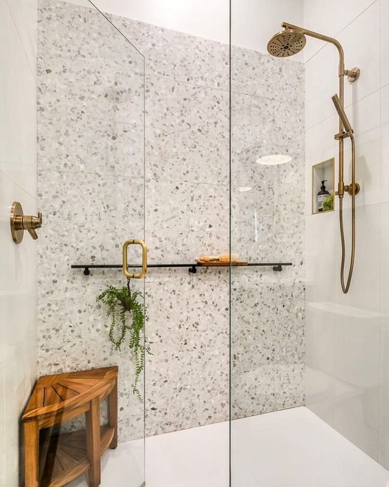 a neutral shower with white tiles and white terrazzo, a niche, a wooden stool, brass fixtures