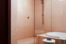 a pink terrazzo bathroom with a shower space, a glass screen, a tub clad with tiles, a marble stool and brass fixtures