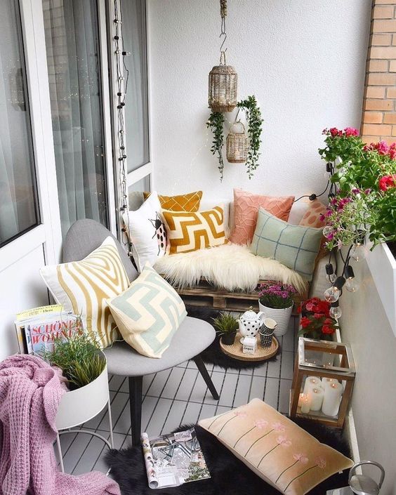 a cool and bright balcony with a pallet sofa with pillows, a chair with pillows, potted bright blooms and lanterns is very inviting