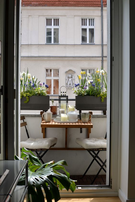 a lovely small balcony with a folding table and chairs, large planters with spring blooms and candle lanterns