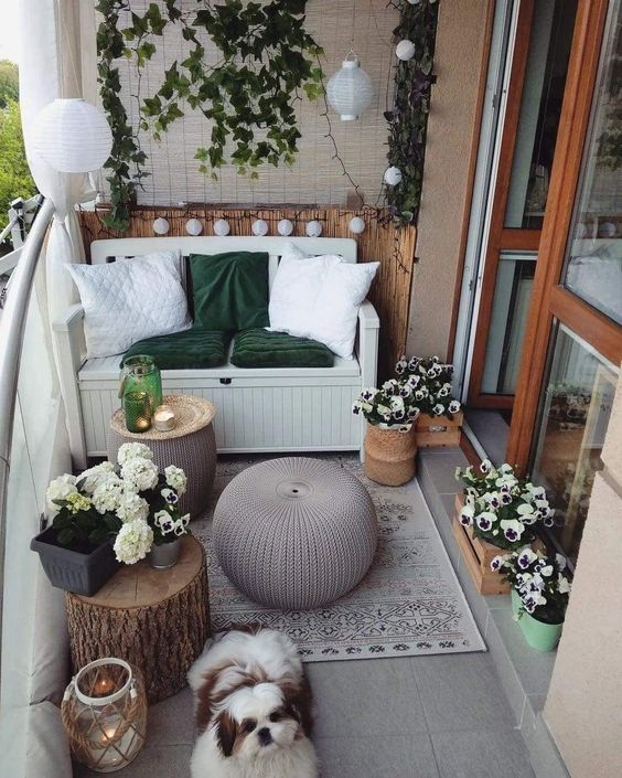a small and lovely balcony with a loveseat and pillows, a stump and some poufs, a rug, greenery and white blooms and lights