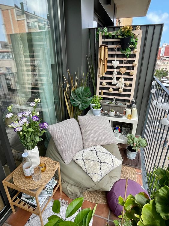 a small and lovely balcony with a potting table with some decor, a beanbag chair with pillows, a side table and blooms and greenery
