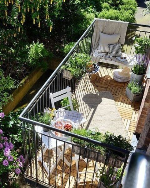a small and welcoming balcony with white folding furniture, potted greenery and blooms and some pillows