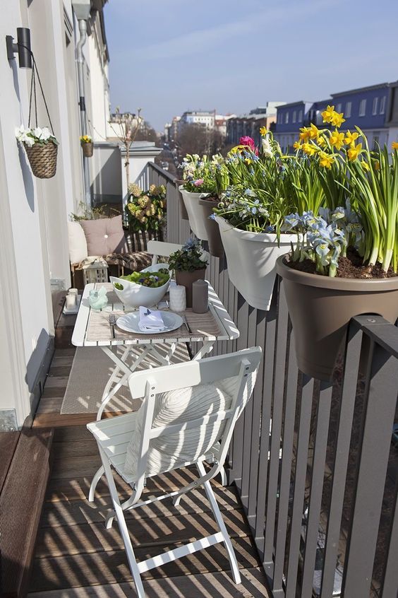 a small white balcony with white folding furniture, a little seat with pillows and railing planters with spring blooms