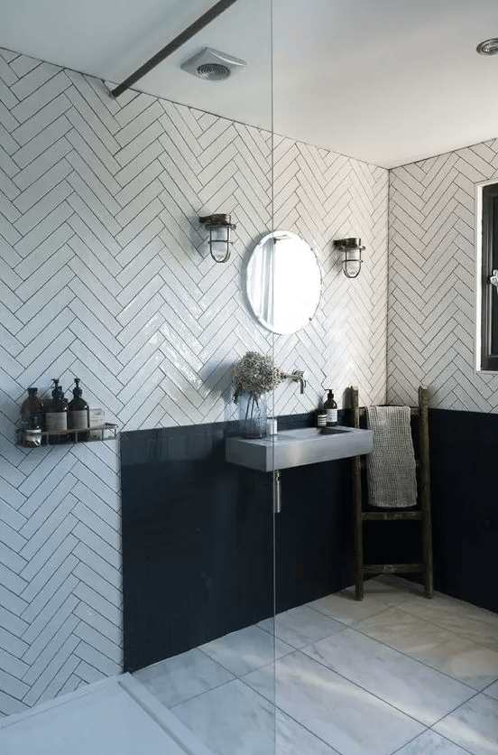 A catchy and chic bathroom with white herringbone tiles, a black backsplash, a concrete wall mounted sink and vintage lamps