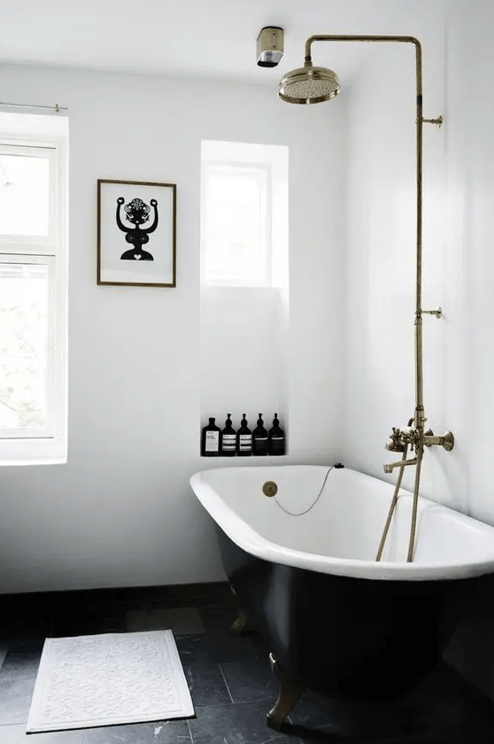 a serene white bathroom with niches for storage, a black clawfoot bathtub, brass fixtures and a fun artwork is cool