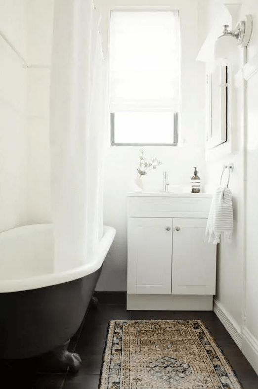 a small boho bathroom with white walls and a dakr-stained floor, a black clawfoot tub, a white vanity and neutral textiles is a chic idea