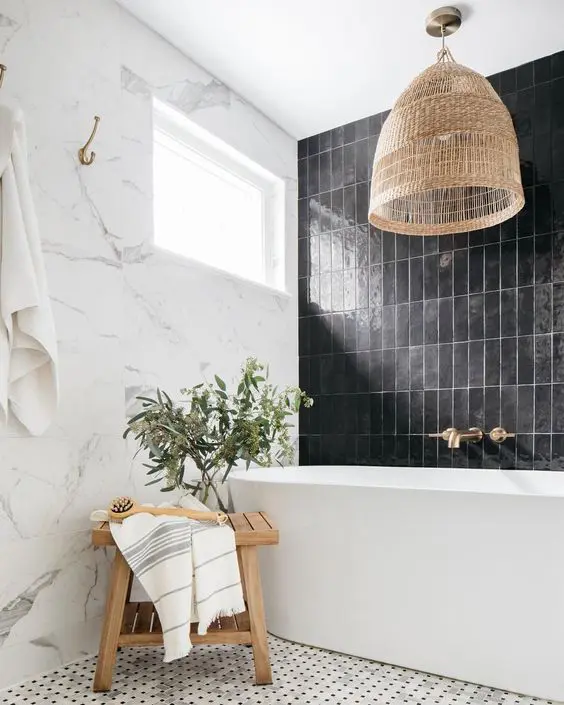 a stylish black and white bathroom with marble and black stacked tiles, a tub, a woven pendant lamp and a stained stool