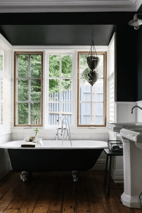 A vintage bathroom with black walls and white planks, a black clawfoot bathtub, free standing sinks and a window