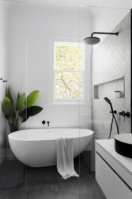 An airy Scandinavian bathroom with grey and white tiles, a floating white vanity, a black sink, a free standing tub and a statement plant