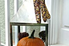 a large lantern filled with pinecones and a large orange pumpkin plus corn on top for a rustic feel