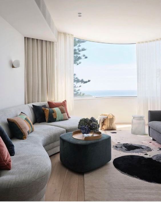 a beautiful curved living room with a curved grey sofa and colorful pillows, a soot pouf, a grey usual sofa and a lovely view