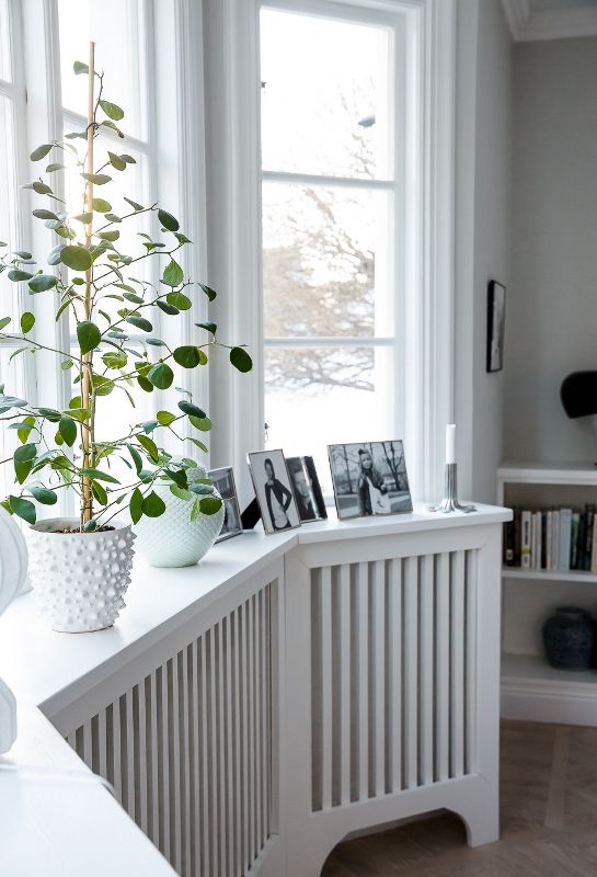 a bow window with radiators covered with planked screens on legs as a windowsill table, with photos and potted plants