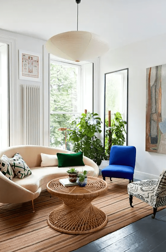 a bright living room with a curved sofa and pillows, a blue and a printed chair, a rattan coffee table, some art and a mirror